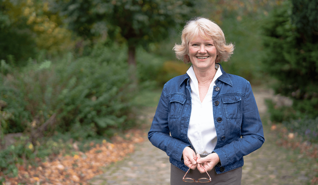 Diese Businessbild brauchst du unbedingt: Jutta Grebing steht in einer blauen Jeansjacke lächelnd draußen im Grünen.