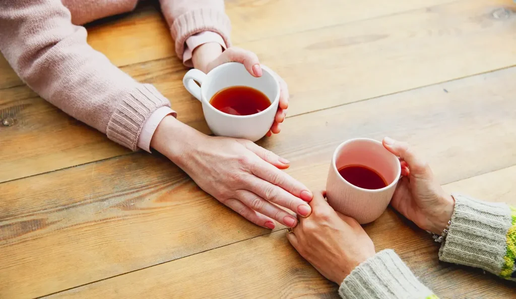 Eine Nahaufnahme von zwei Menschen mit Teebechern, die an einem Holztisch sitzen. Die eine Hand berührt tröstend die Hand von dem anderen.